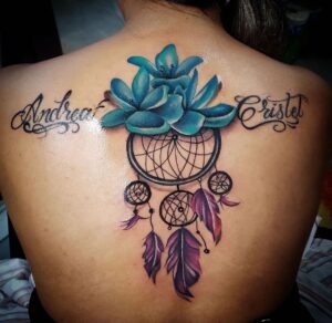 This tattoo features an intricate and colorful dreamcatcher design on the upper back, highlighted by vivid blue flowers at the top. The dreamcatcher itself has carefully detailed webbing and dangling feathers in shades of purple and pink, with smaller dreamcatchers suspended beneath the central one. The tattoo is flanked by elegant script lettering on either side, reading "Andrea" and "Cristel," adding a personal touch to the artwork.