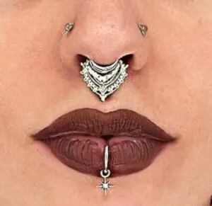 Close-up of a person’s face highlighting facial piercings and makeup. The person has a silver ornate septum ring with intricate details, two symmetrical nostril piercings, and a vertical labret piercing below the bottom lip featuring a dangling star charm. Their lips are painted with dark matte lipstick, emphasizing the piercings.