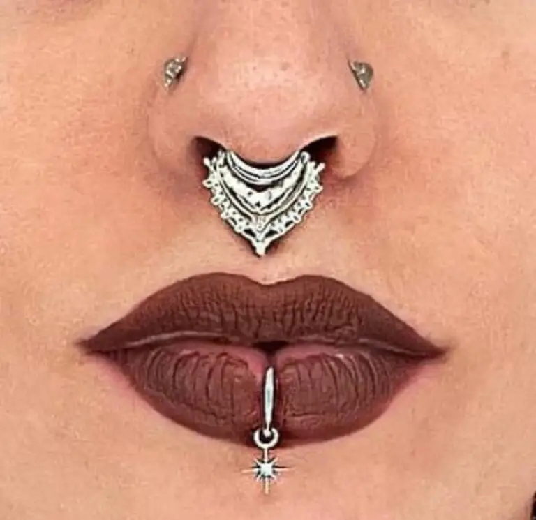 Close-up of a person’s face highlighting facial piercings and makeup. The person has a silver ornate septum ring with intricate details, two symmetrical nostril piercings, and a vertical labret piercing below the bottom lip featuring a dangling star charm. Their lips are painted with dark matte lipstick, emphasizing the piercings.