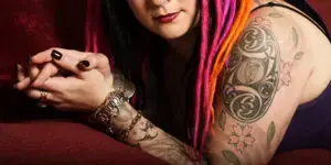 Close-up of a person with colorful dreadlocks, including pink, orange, and black strands, resting on a red couch. Their arm is prominently displayed, showing detailed black and gray tattoos featuring flowers and intricate patterns. They are wearing multiple bracelets, rings, and a dark top. The image highlights their vibrant hairstyle, body art, and jewelry.