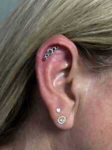 Close-up of a person’s ear with multiple piercings. The upper ear features a curved helix piercing adorned with a row of small, dark gemstones in a decorative setting. The earlobe has two piercings: one with a small diamond stud and another with a gold, circular earring featuring a central pearl surrounded by intricate details. The person's blonde hair frames the ear.