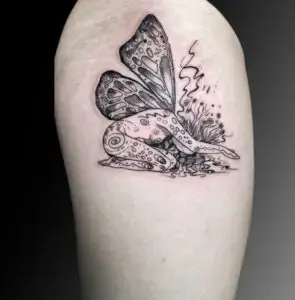 Tattoo of a kneeling fairy with large, intricately detailed butterfly wings on the upper arm. The fairy has a flowing, organic design with swirling patterns on her body, surrounded by abstract plants and delicate linework, creating a whimsical and surreal look.