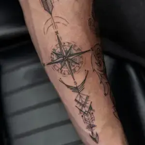 Close-up of a tattoo on a person’s forearm, featuring a detailed black ink compass design with cardinal directions (N, S, E, W) and intricate linework. The tattoo includes an arrow extending through the compass and geometric patterns resembling a stylized arrowhead below it. Additional tattoos, such as an owl and ornamental elements, are partially visible.