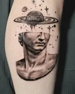 Tattoo of a classical bust with its upper head replaced by a brain surrounded by a ring resembling Saturn's rings. Stars, dots, and a small planet float in the background, adding a cosmic and surreal theme to the design. Shading creates a realistic and three-dimensional effect.