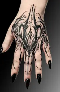 Tattoo on the back of a person’s hand featuring a symmetrical blackwork design with bold lines, intricate details, and sharp, abstract shapes resembling a flame or organic pattern. The tattoo extends down the fingers, complementing the person’s black pointed nails.