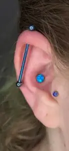 Close-up of a person's ear showcasing multiple piercings. A blue industrial barbell runs diagonally across the upper cartilage, and two piercings feature blue opal gemstones—one in the conch area and one on the tragus. The person's brown hair frames the ear.