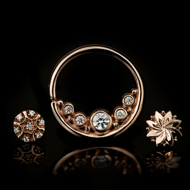 This stunning image showcases rose gold jewelry featuring: A circular barbell adorned with sparkling gems arranged in a crescent design. A floral-inspired stud with intricate detailing. A starburst stud encrusted with dazzling stones. These pieces exude elegance and luxury, ideal for clients seeking elevated styles for piercings like septum, daith, conch, or other curated looks. A circular barbell adorned with sparkling gems arranged in a crescent design. A floral-inspired stud with intricate detailing. A starburst stud encrusted with dazzling stones.