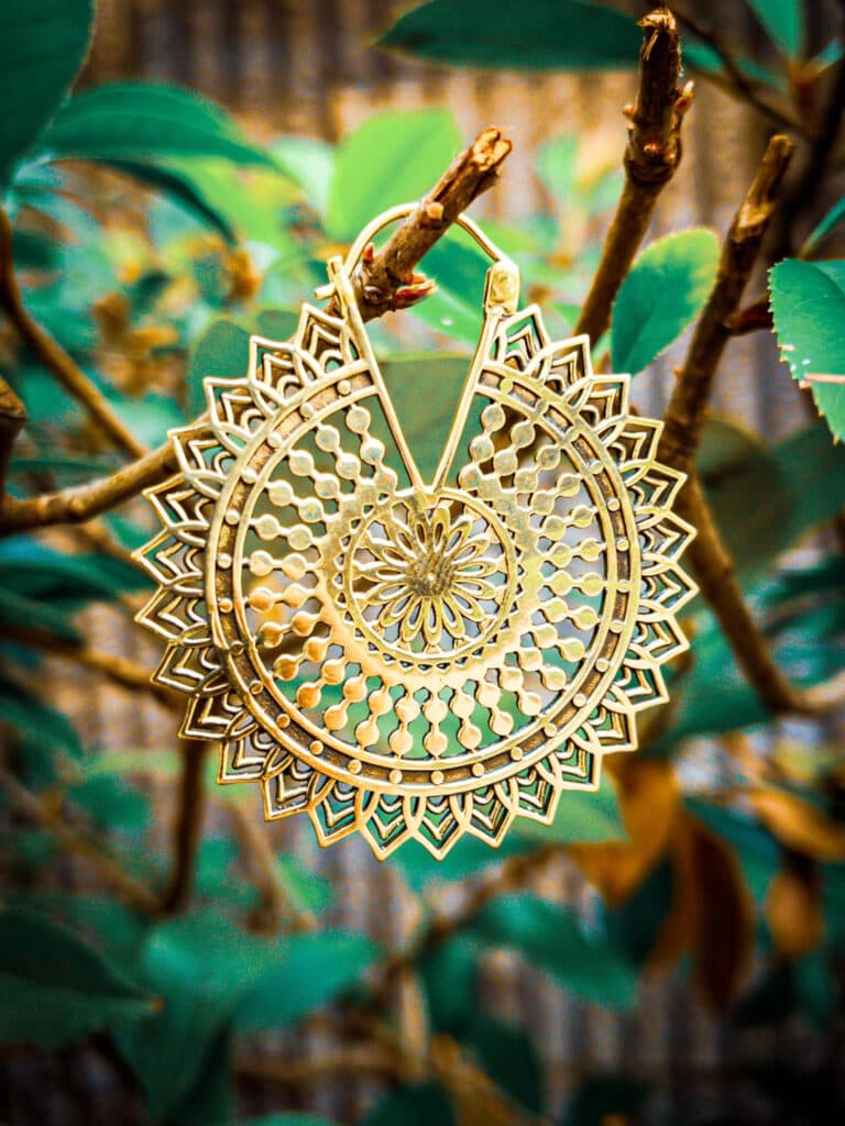 This intricately designed gold mandala hoop earring radiates elegance and craftsmanship. The delicate patterns and circular symmetry make it a stunning statement piece, perfect for a bold yet refined look. The natural backdrop of branches and leaves highlights its bohemian charm.