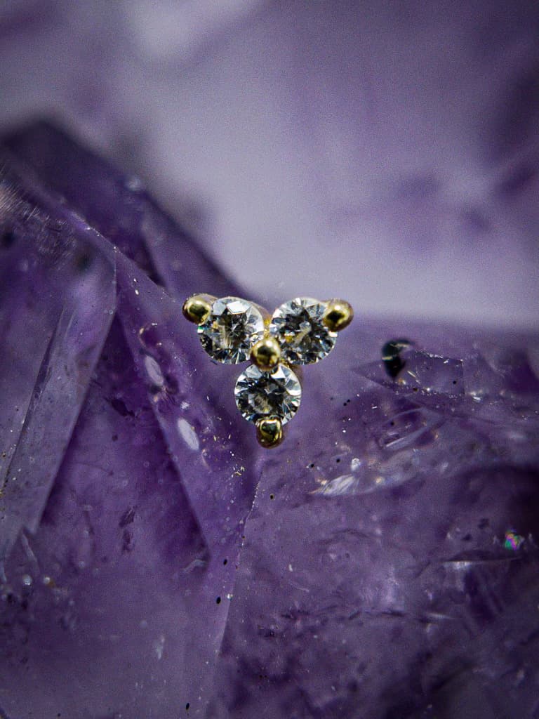 This stunning gold and diamond cluster stud features three brilliant stones arranged in a triangular formation. Set against a striking amethyst crystal backdrop, the piece exudes a perfect balance of elegance and sparkle, making it ideal for luxury piercings.