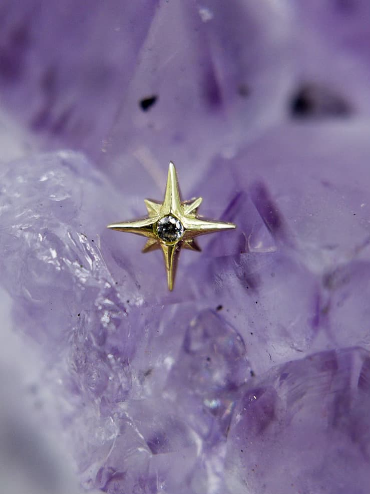 This image features a beautifully crafted gold star-shaped piercing stud with a central gemstone, set against a rich amethyst crystal background. The sharp lines and dazzling stone make this piece stand out, ideal for high-quality ear piercings.