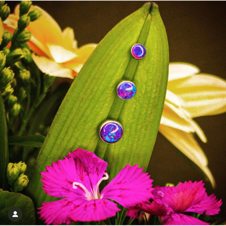 These stunning blue-purple opal studs pop against the lush green leaf and vibrant pink flowers, creating a visually striking, nature-inspired backdrop. Their iridescent glow adds a magical, celestial vibe to the jewelry. Key Highlights: Opal Cabochon Design: A mesmerizing play of blue, purple, and pink hues. Versatile Sizes: Shown in ascending sizes, perfect for curated ear styles or matching sets. Timeless Appeal: Elegant for everyday wear or as statement accents in multiple piercings. Perfect For: Nature-Inspired Jewelry Fans: Compliments earth tones and floral vibes. Curated Ears: Mix-and-match with studs, hoops, or dainty chains. Gift Giving: A thoughtful piece for opal lovers or anyone who adores unique colors.