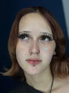 Portrait of a young person with shoulder-length auburn hair, long dramatic eyelashes, and facial piercings. They have a nose ring and a labret piercing below their lip. A small silver star sticker is placed on their cheek, and their gaze is directed upward with a soft expression.