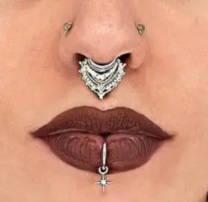 Close-up of a person’s face highlighting facial piercings and makeup. The person has a silver ornate septum ring with intricate details, two symmetrical nostril piercings, and a vertical labret piercing below the bottom lip featuring a dangling star charm. Their lips are painted with dark matte lipstick, emphasizing the piercings.