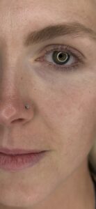 Close-up of a person’s face highlighting a small, silver nose stud with a sparkling gemstone on the left nostril. The person has green eyes, natural eyebrows, and smooth, freckled skin. The image focuses on the details of the eye and piercing.