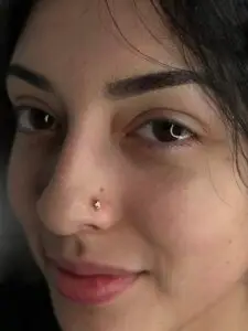 Close-up of a person’s face focusing on a small gold nose stud with a teardrop-shaped design on the left nostril. The person has smooth skin, neatly groomed eyebrows, and natural brown eyes looking softly at the camera. Loose strands of black hair frame the face.