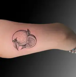 Black and gray tattoo of an orange with leaves and a detailed orange slice, located on the person's inner arm. The design uses dot shading for texture and depth, highlighting the realistic appearance of the fruit and its segments.