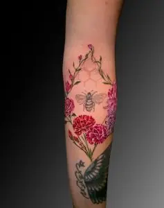 Tattoo on a person's forearm featuring a bee at the center surrounded by a hexagonal honeycomb pattern. The bee is framed by vibrant red and pink flowers with green vines forming a symmetrical arch. The design combines realistic shading with delicate linework for a nature-inspired aesthetic.