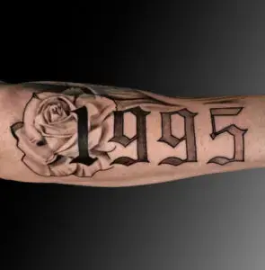 Black and gray tattoo of the year "1995" in bold, Gothic-style lettering on a person's forearm. Behind the numbers, a realistic rose is shaded delicately, creating a soft contrast with the sharp, structured text. The tattoo spans the length of the forearm.