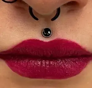 Close-up of a person’s lips painted with bold red lipstick. Above the lips, there’s a black vertical labret piercing with a shiny black gemstone, and a black horseshoe-shaped septum ring. The piercings and makeup create a striking, edgy look.