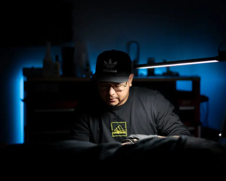 A tattoo artist wearing a black Adidas cap and black shirt works under a focused lamp in a dimly lit studio. The room is illuminated by blue ambient lighting, highlighting the artist’s concentration as they work on a client. Tools and supplies are visible in the background on shelves.