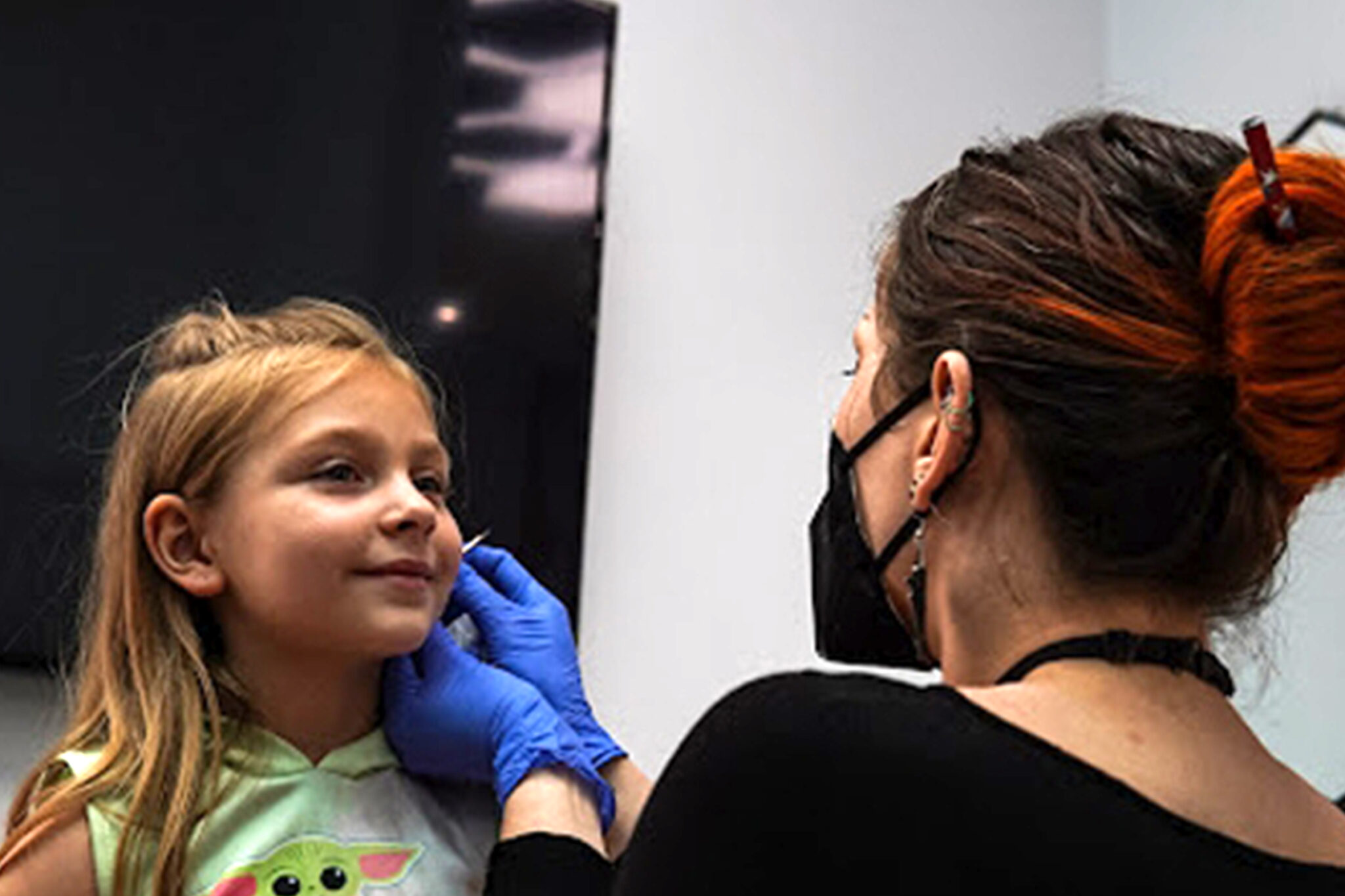 This image captures a child getting an ear piercing at a studio. The professional piercer, wearing gloves and a mask, demonstrates proper hygiene and care during the process. The child looks calm and focused, highlighting a positive experience.