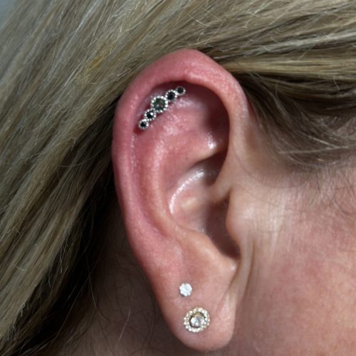 Close-up of a person’s ear with multiple piercings. The upper ear features a curved helix piercing adorned with a row of small, dark gemstones in a decorative setting. The earlobe has two piercings: one with a small diamond stud and another with a gold, circular earring featuring a central pearl surrounded by intricate details. The person's blonde hair frames the ear.