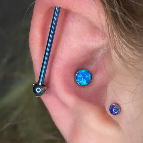 Close-up of a person's ear showcasing multiple piercings. A blue industrial barbell runs diagonally across the upper cartilage, and two piercings feature blue opal gemstones—one in the conch area and one on the tragus. The person's brown hair frames the ear.