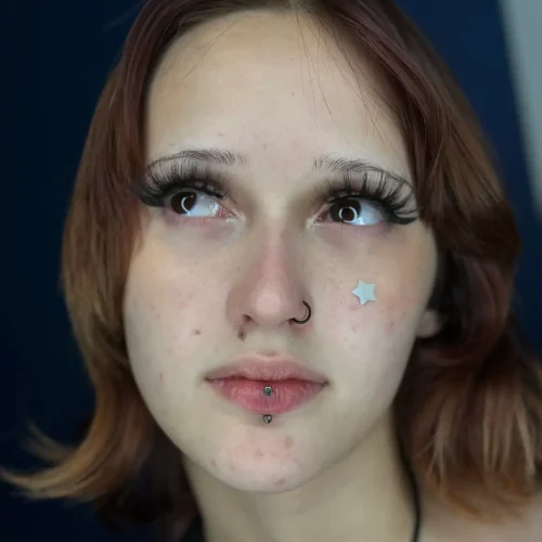 Portrait of a young person with shoulder-length auburn hair, long dramatic eyelashes, and facial piercings. They have a nose ring and a labret piercing below their lip. A small silver star sticker is placed on their cheek, and their gaze is directed upward with a soft expression.