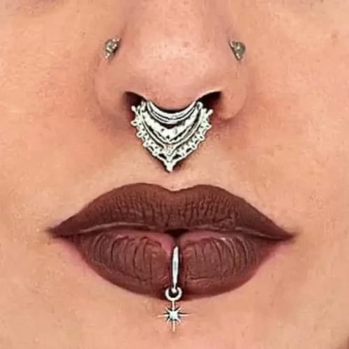 Close-up of a person’s face highlighting facial piercings and makeup. The person has a silver ornate septum ring with intricate details, two symmetrical nostril piercings, and a vertical labret piercing below the bottom lip featuring a dangling star charm. Their lips are painted with dark matte lipstick, emphasizing the piercings.