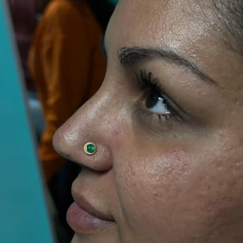 Close-up of a person’s face featuring a gold nose stud with a vibrant green gemstone on the left nostril. The individual has smooth skin, defined eyebrows, and long eyelashes, with their gaze directed upward. A blurred background reveals another person wearing an orange shirt.
